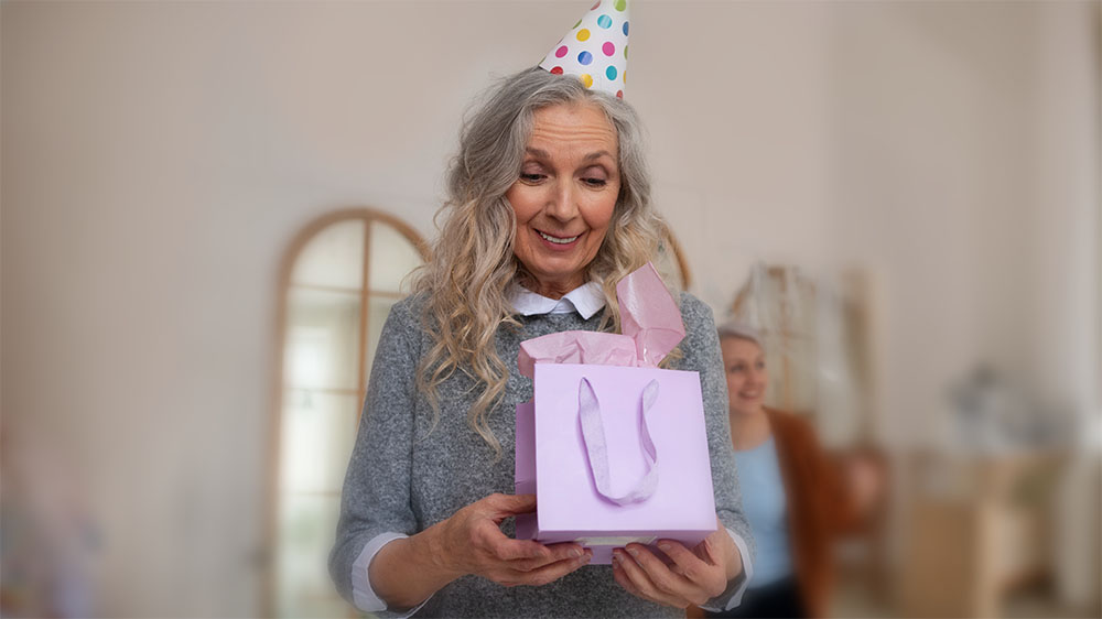 Mulher mais velha sorrindo enquanto segura uma sacola de presente lilás em uma festa de aniversário, usando um chapéu colorido de festa.