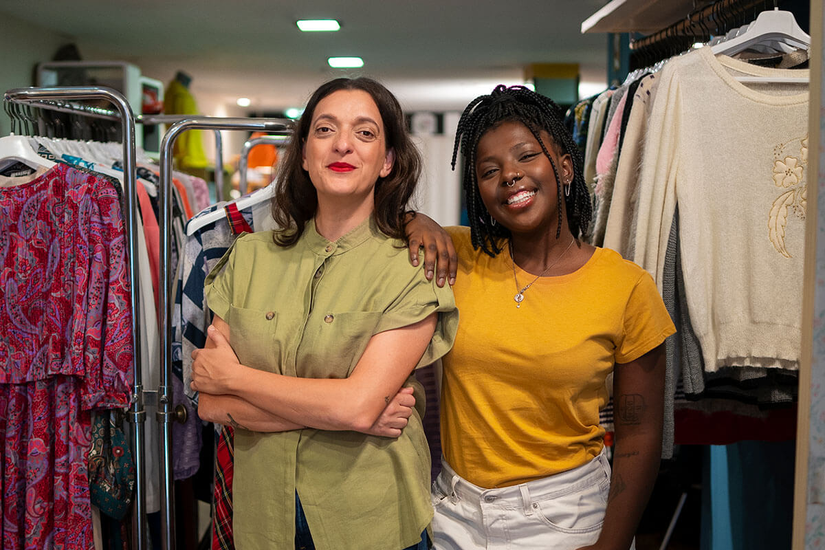 Duas mulheres sorrindo em uma loja de roupas, ambas posicionadas entre cabides de roupas femininas. A mulher à esquerda, vestindo uma camisa verde claro, está com os braços cruzados e uma expressão confiante. A mulher à direita, com uma camiseta amarela, está com o braço apoiado no ombro da outra e também exibe um sorriso.