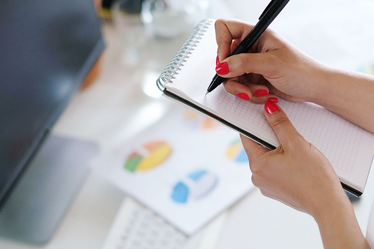 Mãos femininas com unhas pintadas de vermelho segurando um caderno de anotações e escrevendo com uma caneta preta. Ao fundo, um computador e gráficos coloridos desfocados sobre a mesa.